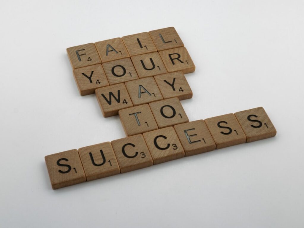 Wooden Scrabble tiles spelling "FAIL YOUR WAY TO SUCCESS" on a white surface.